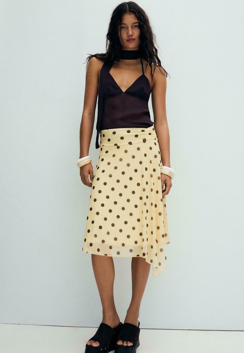 Femme aux longs cheveux bruns portant un haut noir sans manches, une jupe midi jaune à pois marron, des bracelets blancs et des sandales plates-formes noires.