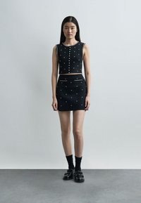 Jeune femme portant un crop top noir sans manches et une jupe courte assortie avec de petits motifs blancs, des chaussettes noires et des mocassins noirs, debout contre un mur uni.