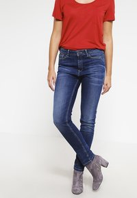 Jeans skiny de color azul oscuro con una cintura de altura media, detalles desgastados y diseño de cinco bolsillos, combinados con una camisa corta de manga roja y botas grises hasta el tobillo.