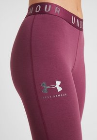 Bordowe legginsy z szerokim elastycznym pasem oznaczonym napisem "UNDER ARMOUR", z srebrnym logo na lewej udzie i gładką fakturą.