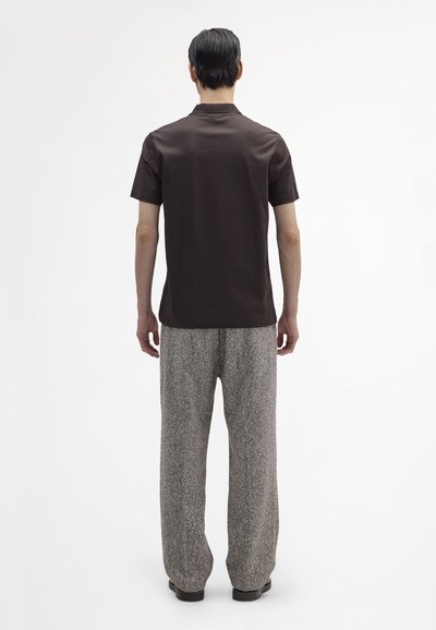 Brązowa koszula z krótkim rękawem w połączeniu z szarymi spodniami o fakturowanym wzorze, luźny krój, gładki kołnierz i płaskie buty. Widok z tyłu.
