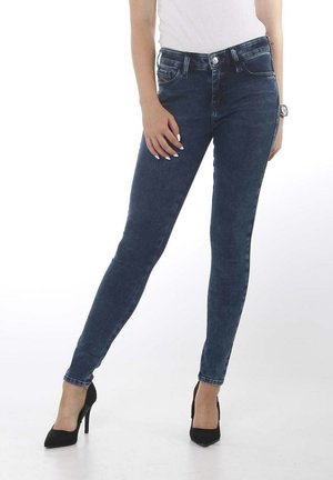 Jeans ajustados de color azul oscuro, confeccionados en denim, con bolsillos, cierre de botón y un ligero desgaste, combinados con zapatos de tacón alto negros.