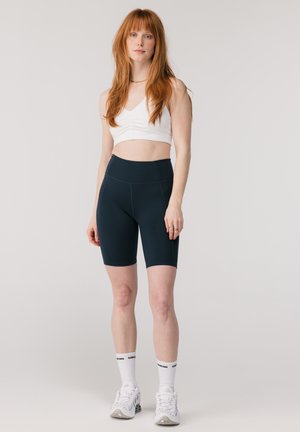 Femme aux longs cheveux roux portant un crop top blanc, un short cycliste taille haute noir, des chaussettes blanches et des baskets blanches, debout devant un fond uni.