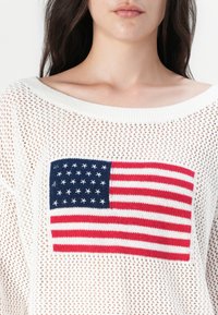 Biely pletený sveter s veľkým vyšitým dizajnom americkej vlajky, ktorý má červené a biele pruhy a modré pole s bielymi hviezdami.