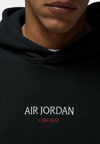 Felpa nera con cappuccio e coulisse, con ricamo bianco "AIR JORDAN" e rosso "CHICAGO" sul petto. Superficie in tessuto texture.