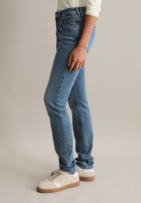 Niebieskie dżinsy o dopasowanym kroju i średnim stanem, z przednimi kieszeniami i gładką fakturą. Połączone z beżowymi sneakersami.