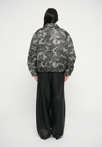 Grijze camouflage bomberjack met volumineuze mouwen en elastische zoom, gecombineerd met brede zwarte leren broeken. Getextureerde stof overal.