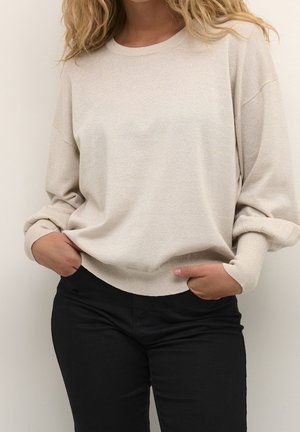 Donna che indossa un maglione beige a maniche lunghe con polsini a costine e pantaloni neri, con le mani infilate nelle tasche dei pantaloni, in piedi davanti a uno sfondo semplice.