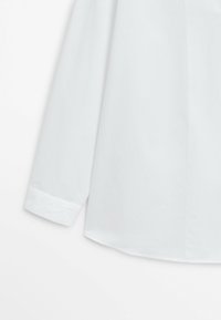 Chemise blanche en coton à manches longues avec poignet boutonné et tissu lisse, présentée de la mi-manche à la taille sur un fond uni.