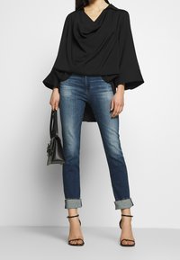 Černý drapovaný top s výstřihem do V, v kombinaci se středně pranými džíny. Texturovaná látka a rozšířené rukávy, doplněné malou černou kabelkou.