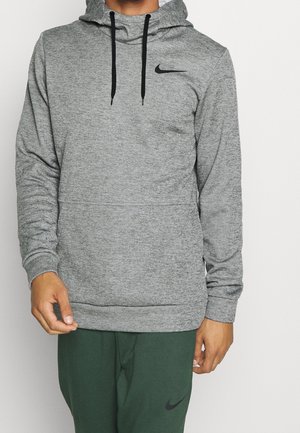 Mężczyzna w szarej bluzie Nike z czarnymi sznurkami i ciemnozielonych spodniach dresowych Nike, stojący na jednolitym jasnym tle.