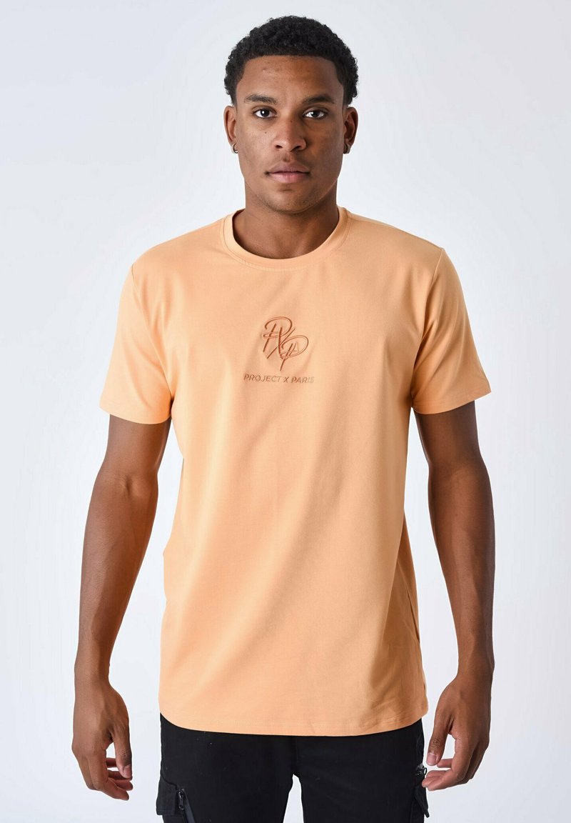 Camiseta de manga corta color durazno, hecha de algodón suave, con un logotipo bordado "PROJECT X PARIS" en el frente. Cuello redondo.