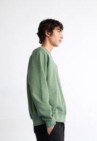 Grüner Baumwollpullover mit gerippten Bündchen und Saum, Rundhalsausschnitt und lässiger Passform, getragen über schwarzen Hosen.