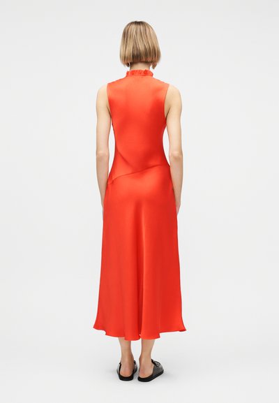 Γυναίκα με κοντά ξανθά μαλλιά, ντυμένη με ένα αμάνικο φωτεινό πορτοκαλί μίντι φόρεμα και μαύρα παπούτσια τύπου slip-on, στεκόμενη με την πλάτη προς την κάμερα σε λευκό φόντο.