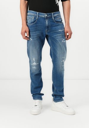 Mann trägt blaue ausgewaschene Jeans und weiße Sneakers, steht vor einem schlichten weißen Hintergrund.