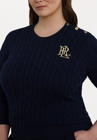 Marineblå kabelstrikket genser med rund hals, med et gullbrodert logo og tre distinkte gullknapper på skulderen.