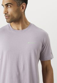 Lavendel bomulls t-skjorte med kort erme og rund hals, med en liten grønn "Champion" logo på brystet og logo på ermet.