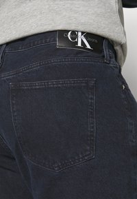 Tmavě modré džíny z denimu s jednou zadní kapsou a černou etiketou Calvin Klein Jeans viditelnou na pase. Měkčená textura a klasický design.