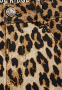 Denim tkanina s uzorkom leopard, u nijansama bež i crne, s žutim šavovima i srebrnim dugmetom označenim "BLUE RIDGE."