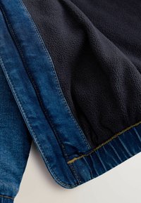 Gros plan sur une veste en denim bleu foncé avec doublure en polaire noire, mettant en valeur les textures du tissu et les détails de couture de la manche et de l'ourlet.