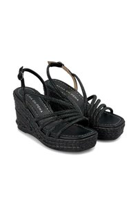 Sandalias de cuña negras hechas de material trenzado con un diseño de tiras brillantes. Cuentan con correas en el tobillo y una suela de cuña texturizada.