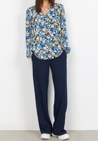 Blommig blus i blå, grön och beige ton med långa ärmar och spetskrage, kombinerad med marinblå vida byxor och vita sneaks.