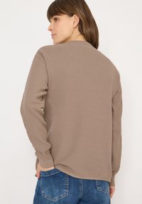 Brązowa prążkowana bluzka z długim rękawem, okrągłym dekoltem i luźnym krojem. Płynnie łączy się z casualowymi niebieskimi dżinsami.