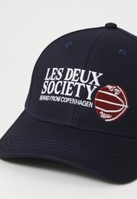 Tengerészkék sapka hímzett "LES DEUX SOCIETY" felirattal és vörös kosárlabda grafikával. Görbített karimával és texturált anyaggal rendelkezik.
