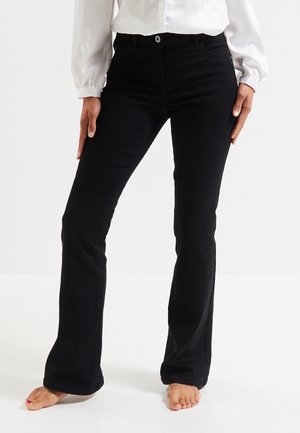 Kobieta ubrana w czarne spodnie bootcut i białą bluzkę z długim rękawem, stojąca boso na białej podłodze na tle jednolitego tła.