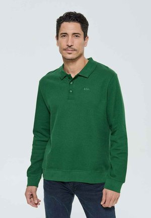 Chemise polo verte à manches longues avec une texture côtelée, patte de boutonnage à trois boutons et logo brodé sur la poitrine, accompagnée de jeans foncés.
