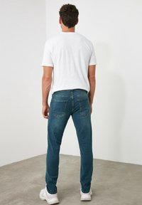 Jeans de mezclilla azul con corte ajustado, detalles de costura visibles y dos bolsillos traseros. Combinados con una camiseta blanca lisa y calzado blanco.
