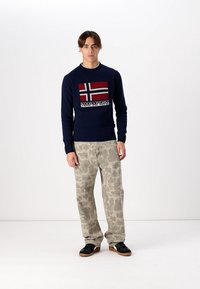 Marineblauer Strickpullover mit einem rot-weißen und schwarzen norwegischen Flaggenmuster, kombiniert mit hellgrauen Tarnhosen und schwarzen Sneakers.