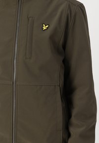 Oliven grønn zip-up jakke laget av vannavstøtende stoff, med en subtil brodert logo og lommer på sidene.