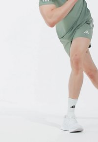 Conjunto atlético que incluye una camiseta verde clara y pantalones cortos, calcetas blancas con logo negro y zapatillas blancas. Tejido suave, diseño ajustado y pantalones cortos hasta la rodilla.