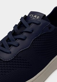 Baskets bleu marine en tissu mesh texturé, accents en cuir, lacets épais et semelle blanche, avec le logo "CLAE" sur la languette.