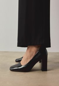 Czarne lakierowane czółenka na słupku z kwadratowym noskiem. Charakteryzują się gładką fakturą i subtelnym wzorem przypominającym skórę krokodyla na powierzchni.