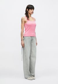 Top de tirantes rosa con textura acanalada combinado con vaqueros de pierna ancha en gris claro. Los vaqueros tienen un detalle de cordón. La modelo está de pie sobre un fondo neutro.