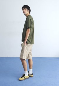 Camiseta de manga corta verde oliva combinada con shorts beige, calcetines blancos y zapatillas amarillas y negras, con un fondo azul claro.