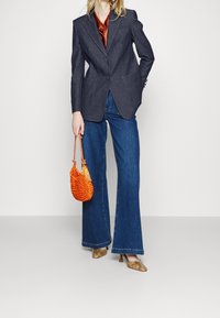 Tummansininen farkkutakki, jossa kaksi etutaskua, yhdistetty leveälahkeisiin sinisiin farkkuihin. Pitelee oranssia punottua käsilaukkua; jalassa kultakuvioidut korkokengät.