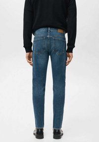 Kapeat siniset farkut, joissa on keskikorkea vyötärö, takataskut ja ruskea nahkainen merkki vyötäröllä. Yhdistetty mustaan neuleeseen ja kenkiin.