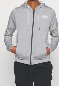 Persona con una sudadera con cremallera de color gris claro y el logo de "The North Face" en el pecho, con las manos en los bolsillos frontales, combinada con pantalones oscuros y texturizados.