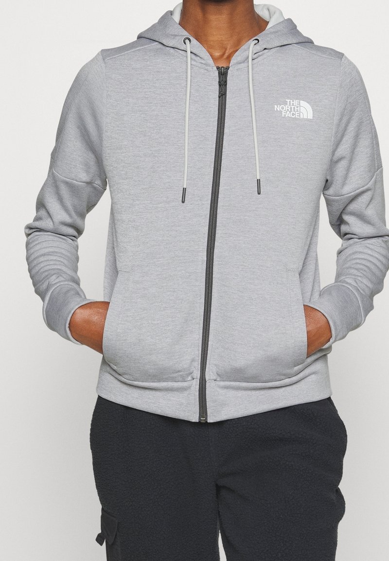 Persona con una sudadera con cremallera de color gris claro y el logo de "The North Face" en el pecho, con las manos en los bolsillos frontales, combinada con pantalones oscuros y texturizados.