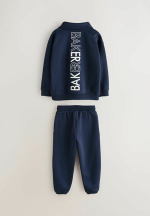 Tumši zils sporta tērps ar kapuci, kas aiztaisāms ar rāvējslēdzēju, un uz muguras vertikāla uzraksta "BAKERY". Bikses ar elastīgu jostu un apkaklīti pie potītēm.