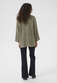 Olive green, textured, long-sleeve shirt with a relaxed fit, paired with dark blue, flared jeans, seen from the back against a neutral background.