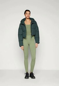 Grøn pufferjakke med hætte over en tætsiddende olivenfarvet jumpsuit. Outfittet har en glat tekstur og er kombineret med sorte sneakers.