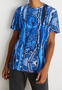 Person klädd i en kortärmad blå mönstrad skjorta med abstrakta vertikala mönster och mörkgrå byxor mot en enfärgad bakgrund.