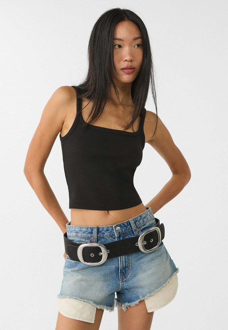 Musta lyhyt cropped-malli tank top, jossa on neliön muotoinen pääntie, yhdistettynä kulutettuihin farkkushortseihin ja leveään mustaan vyöhön, jossa on hopeiset soljet.