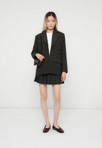 Donker geruite oversized blazer met notched lapels, witte top, geplooide zwarte minirok en glanzende kastanjebruine instapschoenen. Kleine zwarte handtas in de hand.