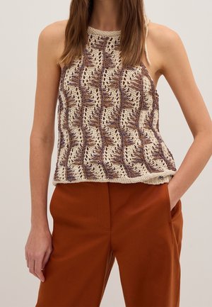 Femme portant un haut en maille sans manches avec des motifs beige, marron et violet, associé à un pantalon couleur rouille, debout sur un fond uni.