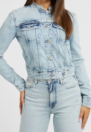 Femme portant une veste en jean bleu clair avec des boutons argentés et un jean taille haute assorti, main posée sur la hanche.
